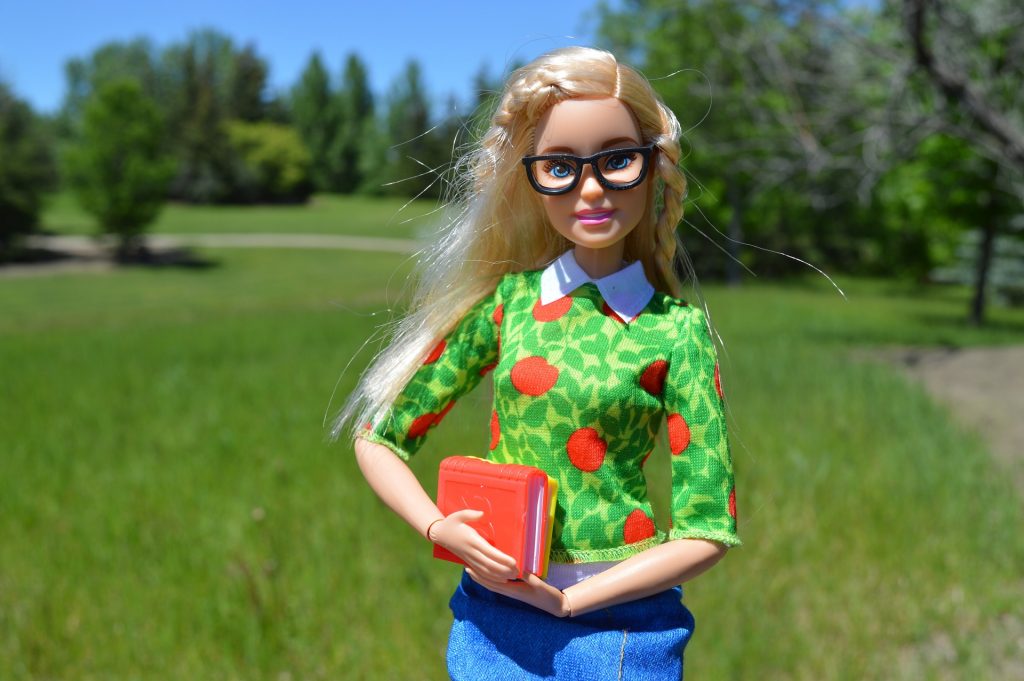Barbie on a walking path, wearing glasses and holding a school book. 